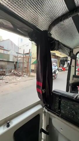 A video shot from inside a vehicle, likely a tuk-tuk, shows a street scene. The interior of the vehicle has a silver insulated ceiling and a dark curtain with red stripes hanging down. Outside the vehicle, a street is visible with buildings on either side. One building appears to be under construction or renovation, with debris and scaffolding. A motorcycle is seen driving down the street, and other vehicles and people are visible in the background. The overall impression is of a bustling urban environment.