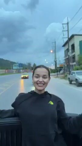 A young woman is riding in the back of a pickup truck on a road. She is wearing a black hoodie with a yellow logo on the left breast. The sky is overcast with dark clouds, suggesting a cloudy day or impending rain. The road has a yellow line and two lanes. Other vehicles, including a yellow sports car and several motorcycles, are visible in the distance and moving along the road. The woman is smiling and appears to be enjoying the ride, waving her arms and striking poses. Buildings and trees line the side of the road, and power lines run alongside the road.