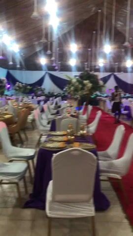 The video shows the interior of a decorated event space, likely for a wedding or celebration. The room has a high ceiling with exposed beams and is lit by overhead lighting. Tables are set with white chairs, gold tablecloths, and purple accents. A red carpet runs down the center of the room, and white and purple drapes adorn the walls. There's a table covered with a red cloth, set with golden serving dishes and plates of fruit. Some people are moving around in the background. The camera movement is quick and somewhat shaky. There is a air conditioner visible.