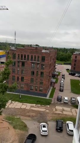 The video begins with a view of a multi-story brick apartment building with many windows, parked cars in a lot in front, and a lone tree with green leaves to the left. The camera then pans to the right, showing a construction crane, several unfinished buildings, a large yellow and blue building in the distance, and more construction materials. Further panning reveals a landscape with trees, a path, and more construction supplies. Finally, the video shows a dense forest and some storage structures, with the sky appearing overcast.