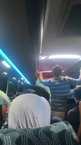 The video shows the interior of a bus with passengers. The perspective is from a seated position, looking forward. A man in a striped shirt is standing in the aisle, holding onto the overhead handrails. He is facing away from the camera and appears to be reaching upwards. Blue LED lights are visible along the side of the bus interior. Other passengers are seated or standing, with their backs to the camera. The overall lighting suggests it is either nighttime or an indoor environment.