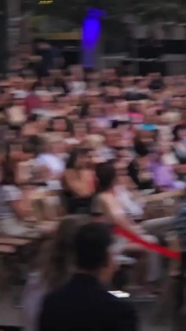 A group of people are seated outdoors, facing a raised platform. A person wearing a black t-shirt and blue jeans walks onto the platform from the left. The person then walks to the right side of the platform and turns to face the audience. Another person, partially visible on the right side of the platform, appears to be holding a microphone or similar device. The environment includes trees, water, and a stone structure in the background. There are also speakers and lighting equipment present on the platform.