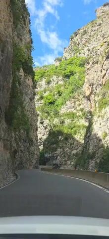 The video shows a car driving on a narrow, winding road through a deep gorge. The walls of the gorge are steep, rocky, and covered in patches of green vegetation. The road is paved and has a concrete barrier on one side. The sky above is blue with some white clouds.