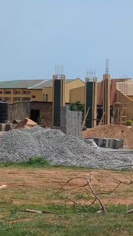 The video depicts a construction site on a sunny day. In the foreground is a pile of grey gravel, and some sparse vegetation on reddish soil. Behind the gravel are stacks of grey concrete blocks, piles of sand, and a black water tank. Further back is a construction site with vertical supports made of wood and steel rebar. Several workers are visible within the structure, seemingly engaged in construction activities. A man wearing a white shirt and black pants walks across the field in the direction of the construction. In the background are several buildings with light-colored walls.