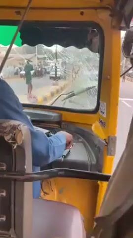 The video is taken from inside a yellow auto-rickshaw, also known as a tuk-tuk. The driver, wearing a blue long-sleeved shirt, is at the wheel. The windshield is severely cracked. Outside, the road is visible with other vehicles and pedestrians. The rickshaw starts to move forward.
