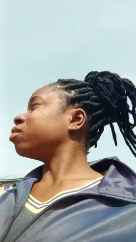 A young woman with dark skin and dark hair in a twisted updo stands outside under a light blue sky. She is wearing a blue and yellow jacket. She looks up and around and then down and straight ahead into the camera. She has a serious expression on her face. The wind is blowing, making the background blurry.