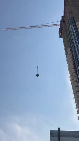 The video shows a construction crane lifting a heavy object. The object appears to be a piece of machinery with wheels, possibly a generator or a compressor. The crane is positioned next to a tall building under construction. The sky is clear and blue. The video focuses on the object being lifted, showing it hanging suspended in the air.