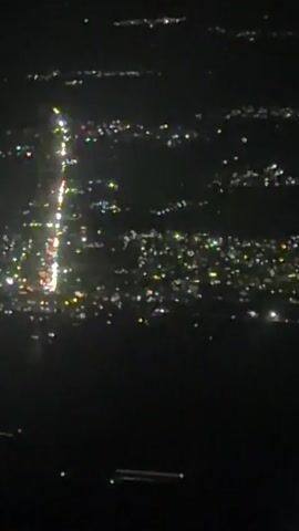 The video shows an aerial view of a city at night from an airplane window. The city lights are visible as a scattering of bright dots and streaks against the dark landscape. A prominent stretch of bright lights, likely a road or highway, runs through the center of the frame. The wing of the airplane, painted a dark blue, is visible in the lower portion of the frame, with a portion of the engine nacelle also visible. The view is slightly distorted, as expected from a window.