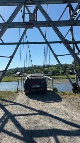 A dark-colored car is slowly driving across a narrow, wooden suspension bridge. The car is the primary subject, seen from behind as it moves towards the center of the bridge. The bridge itself is constructed with metal beams and cables, supporting wooden planks that form the road surface.

The scene is set in a rural, mountainous area. Lush green hills and trees are visible on either side of a river that flows beneath the bridge. Several houses with red roofs are scattered across the landscape, suggesting a small village or settlement. The sky is clear and blue, indicating a sunny day. The time of day appears to be midday or early afternoon due to the bright sunlight and the shadows cast on the bridge and the road leading up to it.

The car's license plate is visible and reads "M193PA 142". There are also some stickers on the rear window of the car. The overall impression is one of a deliberate and perhaps cautious journey across a somewhat precarious bridge in a scenic, natural setting. The activity is clearly the car traversing the bridge. There are no people visible in or around the car, adding a sense of quiet progression to the scene.