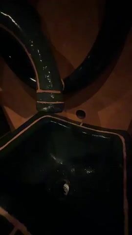 A close-up video captures a unique sink and faucet setup in a dimly lit environment. A person's hand, adorned with a ring, is extended into a dark green, rectangular sink basin with lighter, possibly orange or brown, trim. Above the sink, a long, winding dark green pipe with matching trim serves as the faucet, curving down towards the basin. As the hand is placed under the faucet, a thin stream of water begins to flow from the pipe's end into the palm of the hand and then into the sink. The background wall is a warm, earthy brown or terracotta color. Other dark green, similarly trimmed, sculptural elements are visible on the wall, suggesting a decorative or artistic theme throughout the space. The overall impression is one of a uniquely designed washroom or public restroom facility.