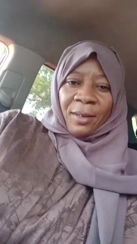 A woman is seated in the driver's seat of a car, facing the camera. She is wearing a mauve hijab and a patterned mauve blouse. She smiles at the camera, revealing a few teeth. In the background, out the car window, a tree is visible. Music is playing in the background.