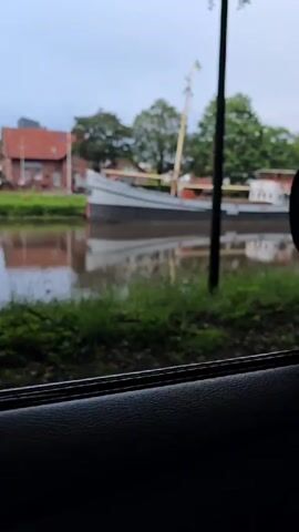 The video shows a view from inside a vehicle looking out a window at a canal. On the canal, there are several boats. The foreground shows the interior of the car, including the door panel and side mirror. In the middle ground, there is a body of water, which is a canal. Along the edge of the canal, there is greenery and some trees. Further in the background, there are buildings and trees. The sky is overcast. The boats on the canal appear to be barges or cargo ships. One boat is grey and white with a mast and a yellow and blue flag. Another boat is longer and has an orange and white section. The water reflects the boats and the sky. The trees on the right side of the frame are large and leafy, partially obscuring the view. There is a black pole in the center of the frame, possibly a lamppost, that is partially visible. The overall scene suggests an overcast day in a town or city with a canal running through it.