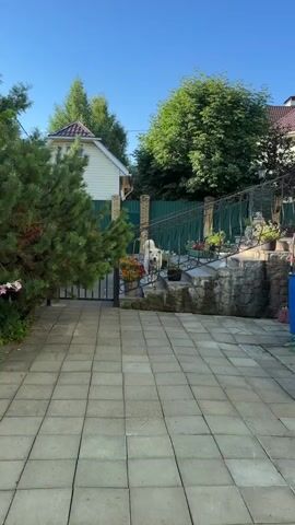 A white dog stands on a stone staircase outdoors. The staircase has a metal railing and potted plants. Behind the staircase is a green fence and a small building with a white exterior and a beige roof. To the left, there is a large green pine tree and a metal gate. The ground in front of the staircase is paved with square gray tiles. The sky is clear and blue.