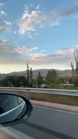 The video shows the view from a car window driving along a highway. The sky is visible, with a mix of blue and white clouds. Trees line the side of the road, and a mountain range is visible in the distance. A wind turbine is faintly visible among the mountains. The car's side mirror is visible in the foreground. The camera appears to be moving with the car.