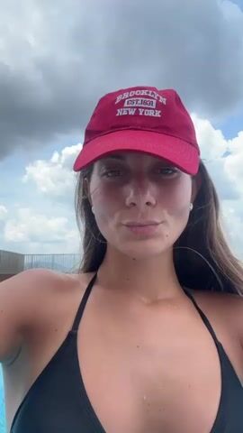 A young adult woman is seen in an outdoor swimming pool under a partially cloudy sky. She is wearing a black halter-neck bikini top and a red baseball cap with white text that reads "BROOKLYN EST 1831 NEW YORK".

Initially, she looks directly at the camera with a calm, neutral expression. She then raises both hands to the back of her head, appearing to adjust her hair or pose, while turning her body slightly to the side. As she turns, she looks over her shoulder at the camera with a subtle, confident smile.

The pool water is clear and blue, with gentle ripples. In the background, beyond a metal railing that lines the edge of the pool deck, distant buildings and structures are visible under a sky featuring both dark gray clouds and patches of bright blue. The scene suggests a rooftop pool setting in an urban or suburban environment, consistent with Ampang Jaya Municipal Council, Malaysia. The lighting is bright but diffused due to the overcast conditions.