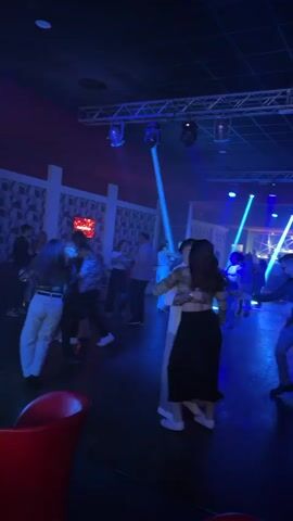 The video depicts a dimly lit nightclub filled with people dancing and socializing. Blue spotlights and other stage lighting fixtures illuminate the scene. There are couples dancing on the dance floor, and others are sitting on red couches along the walls. A metal truss is visible on the ceiling, supporting the stage lighting. The club has a modern design with textured wall panels. Several people are wearing light-colored clothing, while others are in darker attire. Some individuals are also observing the scene from the seating area.