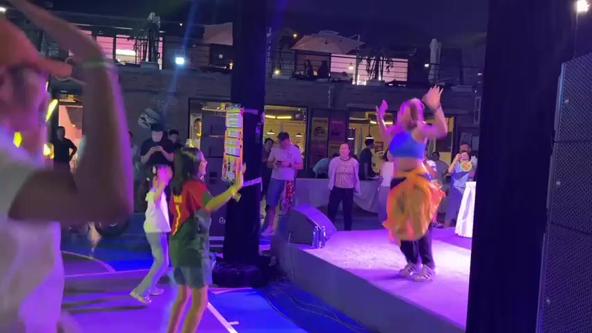 The video shows a lively outdoor event at night. A woman in a blue crop top and black pants with a yellow sash is dancing on a stage. She has her arms raised and appears to be performing a choreographed routine. Several children are dancing on the ground in front of the stage, to the left of the frame. They are wearing casual clothing. In the background, there is a crowd of people watching the performance. Some are standing, while others are seated at tables. The environment appears to be a restaurant or entertainment venue, with brick buildings and string lights visible. There are speakers and stage lighting set up for the performance. The overall atmosphere is festive and energetic.