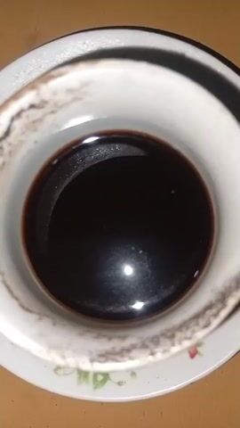 A close-up, overhead shot focuses on a white cup filled with a dark liquid, likely coffee. The cup rests on a saucer with a floral pattern. The rim of the cup appears slightly stained or worn. The lighting in the scene creates a bright reflection on the surface of the liquid. The background is a warm, light brown, possibly a wooden surface. The video appears to be a static shot, with no discernible movement.