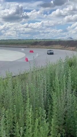A black hatchback is shown driving on a paved track. The car is navigating a corner with orange and white cones placed around the track. In the background, there is a wide expanse of green fields, a body of water, and a cloudy sky. Another white car is visible further down the track. There are also several white rectangular markers and red cones set up along the track. In the distance, a small building with a tower is visible. The foreground is filled with tall, green vegetation. The video appears to capture a moment from a driving event or practice session.