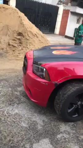 The video shows a red Dodge Charger parked outdoors on a paved surface. The hood of the car is black with flame decals. There is a pile of sand next to the car. A man is standing in the background wearing a green and black shirt. The car has some visible damage on the front passenger side. There is a building and a fence visible in the background. A white vehicle passes by in the background.