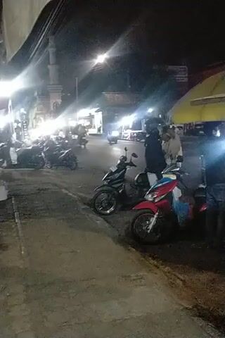 A nighttime street scene with several motorcycles parked on the side of the road. The area appears to be an open-air market or food stall with a yellow and red umbrella providing shade. Bright lights illuminate the area, and people are seen interacting near the motorcycles and the stall. One person is lying on the ground near a motorcycle. The ground is paved and appears to be a street or sidewalk.