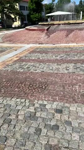 The video pans across a public square on a sunny day. In the center is a large, shallow brick fountain with water jets creating a misty spray. The fountain is surrounded by a paved area with geometric patterns of red, gray, and tan bricks. Beyond the fountain, there is a white open-air structure, possibly a pavilion, and various trees and greenery. In the distance, there are buildings and people can be seen walking. The sky is blue with scattered white clouds.