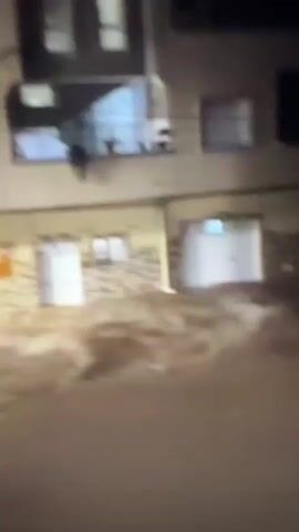 A video shows a street flooded with murky brown water. The water level appears to be at least waist-high in some areas, submerging vehicles and the lower parts of buildings. The buildings have multiple stories, with some showing signs of damage or neglect. There are objects floating in the water, including what look like debris, possibly furniture, and parts of vehicles. The lighting suggests it is nighttime or very dark. The camera is moving, indicating someone is filming from a vehicle or while walking through the flooded area. The sounds in the video include rushing water and what sounds like Arabic speech. The speech seems to express concern or a prayer related to the flood.