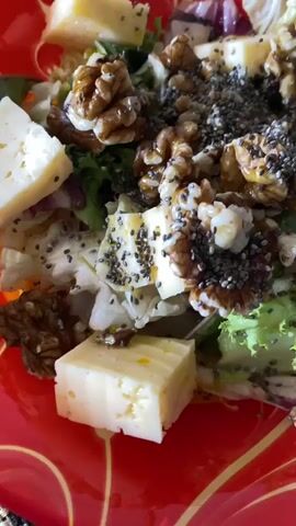 A close-up shot of a salad in a bright red bowl. The salad is composed of mixed greens, cubes of cheese, walnuts, and sprinkled with chia seeds. The camera slowly pans across the bowl, highlighting the texture and arrangement of the ingredients. The lighting appears to be natural, casting a soft glow on the food. The red bowl has a floral pattern around its rim.
