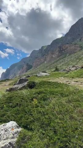 This is a vertical video showcasing a breathtaking mountain landscape in El'tiubiu, Russia. The scene is dominated by majestic, rugged mountains, their slopes partially covered in green vegetation and rocky outcrops. A winding dirt path is visible in the foreground, suggesting a hiking trail. The sky is a vibrant blue, adorned with fluffy white clouds that create dramatic shadows and highlights on the mountainsides. The time of day appears to be daytime, likely late morning or early afternoon, given the bright sunlight. The weather is clear and sunny, with the clouds adding a dynamic element to the otherwise serene atmosphere. The overall impression is one of natural beauty, vastness, and tranquility. There is no visible text in the media file.