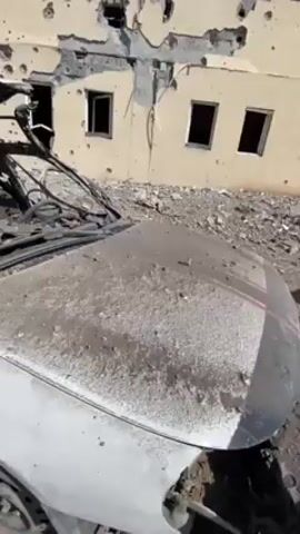 The video shows a destroyed white car in front of a building riddled with bullet holes. The car's windshield is completely shattered, and the interior is filled with debris. The car's seats are visible but heavily damaged. The building behind the car shows significant structural damage from apparent gunfire or explosions. The overall scene suggests a war zone or area with recent heavy conflict. There are no people visible in the video. The camera slowly pans across the car and building to highlight the extensive damage.