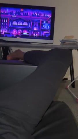The video shows a first-person perspective of someone sitting down, with their legs visible in black sweatpants. In the background, a flat-screen television is displaying a colorful, animated image of a city with purple and pink hues. A table with some papers and other items is positioned behind the TV. The person's hand is visible in the lower left corner of the frame.