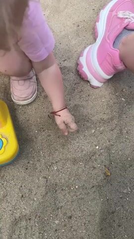 The video shows a close-up view of a young child's arm and hand playing in sand. The child is wearing a pink t-shirt, pink shoes, and a red bracelet on their left wrist. Their hand is covered in sand as they reach down and pick up a small white shell. To the left of the child's arm is a yellow toy bucket. In the upper right corner of the frame, a pink and white sneaker is visible, suggesting another person, possibly an adult, is nearby. The environment is sandy, likely a sandbox or beach. The lighting appears to be natural daylight.