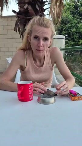 A woman with blonde hair pulled back in a ponytail, wearing a peach-colored tank top, sits at a white table outdoors. Her hands are clasped in front of her, and she is holding a cigarette. To her left is a red mug with the word "LOVE" printed on it multiple times in white. In front of her is a silver ashtray with cigarette butts and ash. To her right is a pack of cigarettes and a pink lighter. She appears to be speaking to the camera. Behind her, a palm tree and greenery are visible, along with a brick wall and a railing.