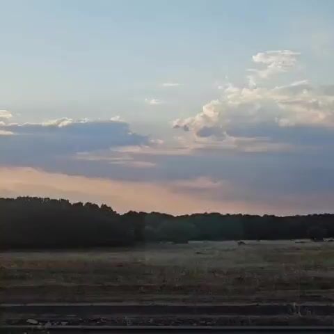 The video shows a time-lapse of a sunset over a rural landscape. The sun is setting behind a line of trees, casting a warm glow across the sky. Clouds are scattered across the sky, and some are illuminated by the sun's rays. A field is visible in the foreground, with a road running along the bottom of the frame. The overall impression is one of peace and tranquility.