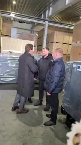 The video shows three men greeting each other in a warehouse-like space. The first man, wearing a long black coat and grey pants, shakes hands with the second man, who is wearing a blue puffer jacket.  The third man, wearing a black puffer jacket, smiles and nods. Large crates, some wrapped in plastic, are stacked in the background. The space has a high ceiling with metal beams and bright lighting.  The floor appears to be concrete.