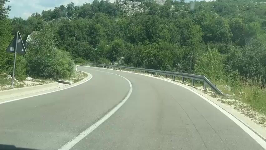 The video shows a view from a moving vehicle on a winding paved road. The road has a white line dividing the lanes. On the right side of the road, there is a metal guardrail. The environment is characterized by a hilly landscape with lush green trees and some rocky outcrops. The sky is visible with white clouds. The road is curving to the left, and the vehicle appears to be driving uphill. There are white marker posts along the side of the road.
