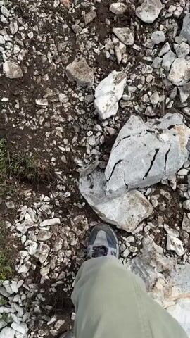 A person's feet wearing a gray and blue hiking shoe and green pants are visible in the foreground of the video. The camera is pointed downwards at a rocky, uneven ground covered with stones and dirt. The terrain appears to be a mountain path. As the video progresses, the camera pans up and to the right, revealing a steep, grassy slope dotted with small yellow flowers. Two figures, presumably people, can be seen ascending the slope in the distance. The sky is overcast with clouds. The video captures a hiking or trekking experience in a mountainous environment.