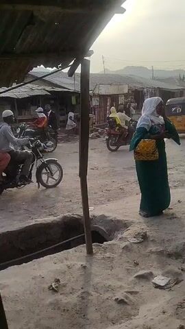 The video shows a street scene in what appears to be a small town or village.  A motorcycle with two passengers drives past, moving from left to right. A woman in a green dress and white headscarf stands on the side of the road, near an open pit. The environment looks dusty, and the buildings are small and possibly constructed with corrugated metal. A makeshift shelter with a tattered metal roof is visible in the foreground, obscuring the view somewhat. There are other people walking in the background, and the lighting indicates it is daytime.
