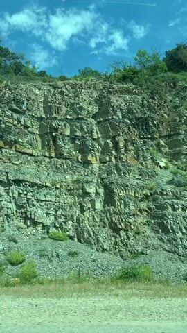 The video shows a rocky hillside with visible rock layers and some scattered vegetation. The camera appears to be in a moving vehicle, as the scenery moves from left to right across the frame. The sky is partly cloudy with patches of blue. In the foreground, there is a narrow strip of green grass and bushes, suggesting a road or path runs alongside the base of the hill.