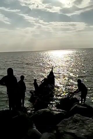 The video shows a small boat being brought to shore by a group of people. The scene is silhouetted against the bright sunlight reflecting off the ocean. Several people stand on the rocky shore, assisting with landing the boat. One person seems to be handling a net. The sea stretches to the horizon under a cloudy sky.