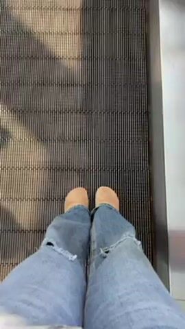 A first-person perspective shot shows a person standing on a moving walkway. The person wears blue jeans with ripped knees and tan-colored shoes. The moving walkway has a textured, metal surface. Sunlight casts shadows across the walkway, creating alternating light and dark stripes. The camera is static and the walkway is moving.