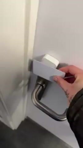 A person is using a white card to open a door with a chrome handle. The person inserts the card between the latch and the door frame, then pulls the handle to open the door. The door is gray with white trim. The floor is a light gray color. The person's hand is visible, and it has a tattoo on it. The person is wearing a black jacket.