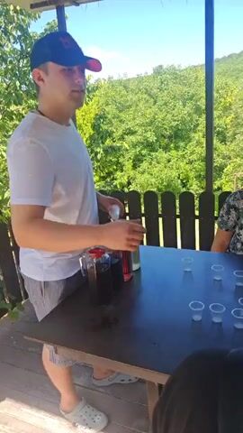 A man wearing a white t-shirt and a baseball cap stands on a wooden deck, holding a small white object and talking on his phone. A table in front of him displays several large glass jars filled with dark red and light-colored liquids, along with a pitcher and several small plastic cups. Behind the table, a wooden fence encloses the area, with lush green trees and a hillside visible in the background under a bright blue sky. To the right of the man, a woman wearing a floral patterned shirt is partially visible. In the background, another woman with glasses and a black top is seated at the table, and further back, a younger woman with dark curly hair and a black t-shirt is also seated. Another person's shoulder and arm, wearing a pink garment, are visible in the foreground on the right. The scene suggests an outdoor gathering or tasting event.