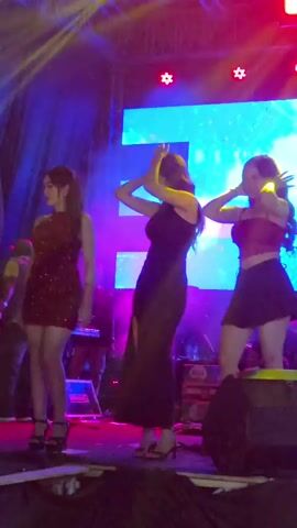 Three young women are dancing on a stage. The woman on the left is wearing a short, sparkly red dress and high heels. The woman in the middle is wearing a long, black dress with a slit on the side and high heels. The woman on the right is wearing a red crop top and a short black skirt, with high heels. They are all illuminated by colorful stage lights, including red, pink, and yellow. In the background, there is a large screen displaying abstract colorful graphics. There are also some musical instruments visible on stage, including what appears to be a keyboard. The overall atmosphere is that of a live performance or concert.