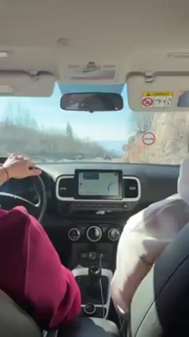 A first-person view from the back seat of a car shows two people driving along a road. The driver wears a maroon sweatshirt, and the passenger wears a light pink hoodie. The car has a navigation system displayed on the dashboard screen, and the driver has both hands on the steering wheel. Outside, the sun is shining, and the scenery consists of trees, rocks, and a distant mountain range. A speed limit sign is visible on the side of the road. The car appears to be driving on a winding road through a mountainous area.
