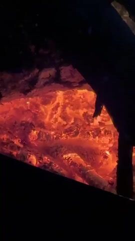 The video shows a close-up view of glowing red embers in a dark environment. The embers are primarily in the center of the frame, with some darker areas suggesting ash and charred wood. The light from the embers illuminates the surrounding darkness. There are no people, animals, or other objects visible. The scene appears to be a fire pit or a fireplace with the fire having burned down to embers.