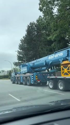 A large blue Sarens brand mobile crane with multiple axles is driving down a paved road. The crane has the company logo visible on its boom. The road appears to be in an industrial or suburban area, with trees and buildings visible in the background. The sky is overcast. The video is shot from a low angle, likely from inside another vehicle.