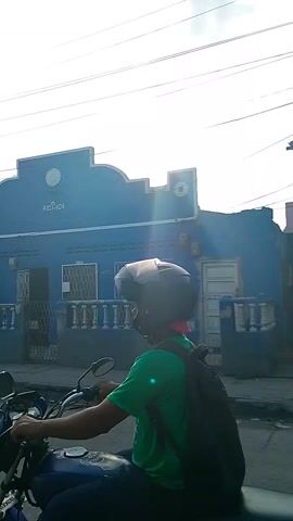 The video starts with a low-angle shot of a blue building with the word "RICARDA" visible on its upper portion. The sky is partly cloudy with visible power lines. A person wearing a helmet and a green shirt is seen riding a motorcycle. The camera then pans to show a yellow car driving down a street lined with buildings and trees. The video ends with a blurry close-up of a dark, undefined object.