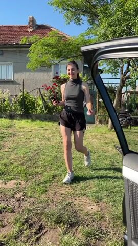 A young woman walks across a grassy, uneven yard in Rogozen, Bulgaria, on a sunny day. She is wearing a dark grey sleeveless turtleneck top, black shorts, and white athletic shoes. Her long dark hair is pulled back, and she holds a dark-colored phone in her left hand. She is smiling and appears to be looking towards her right, possibly at someone or a vehicle partially visible on the far right.

In the background, a house with a red-tiled roof and light grey walls is visible, with white-framed windows. A garden area with green foliage and red roses sits in front of the house, enclosed by a teal-colored metal gate. Trees and other greenery are scattered around the property. The ground shows patches of bare earth and some tire tracks. The clear blue sky indicates bright, pleasant weather. No visible text is present.