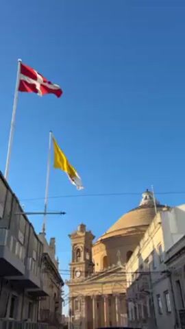 The video shows an external shot of a building with architectural features resembling those of a church. The dome and bell tower are clearly visible. A red and white flag flutters on a pole on the left, and a yellow and white flag hangs from another pole to its right. The sky is clear and blue. Some buildings are visible on the left, with balconies. The recording is taken from street level, looking up towards the building and flags.